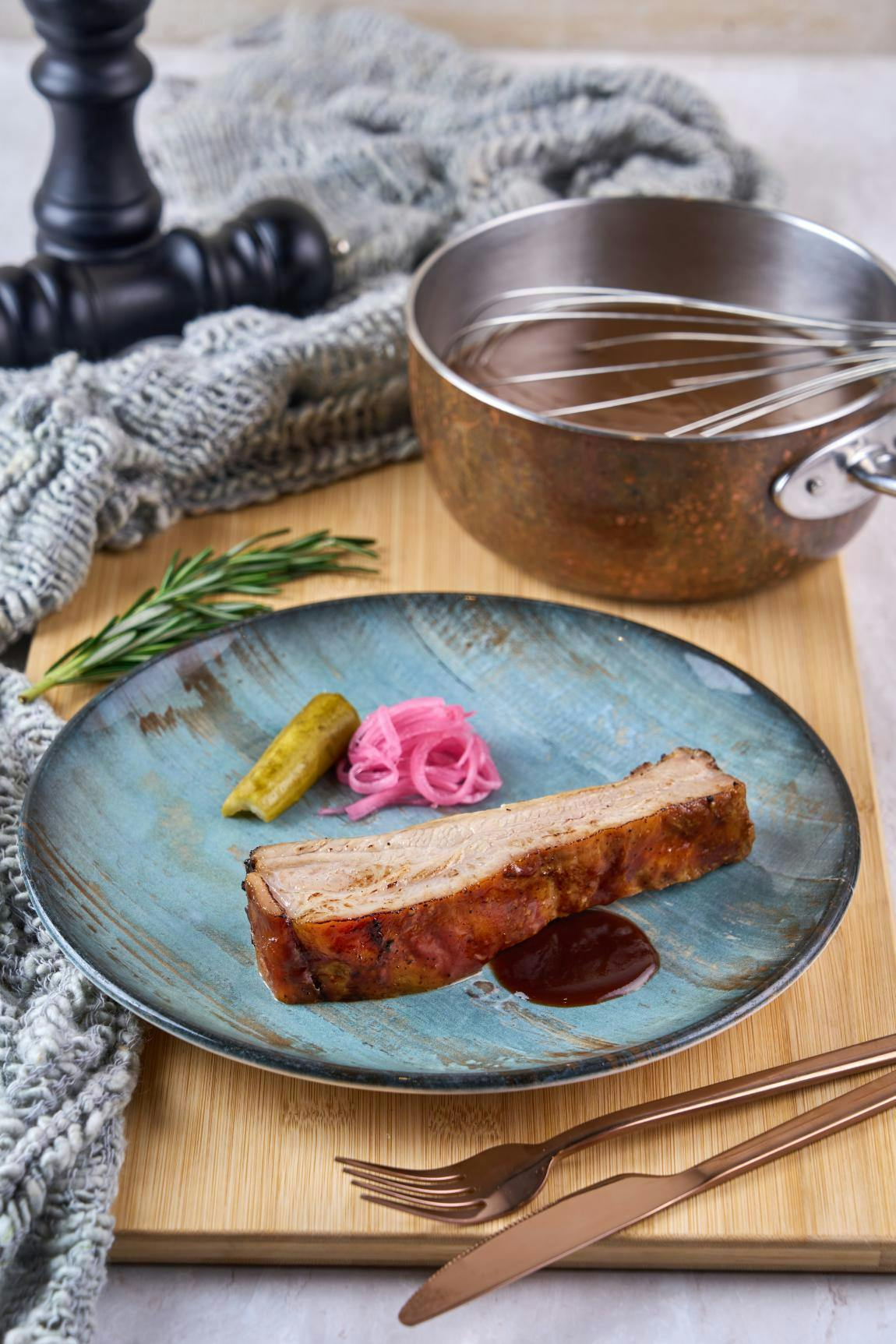 A plated dish with a slice of roasted pork, pickle, pickled onions, and sauce, next to a pot and whisk on a wooden surface.