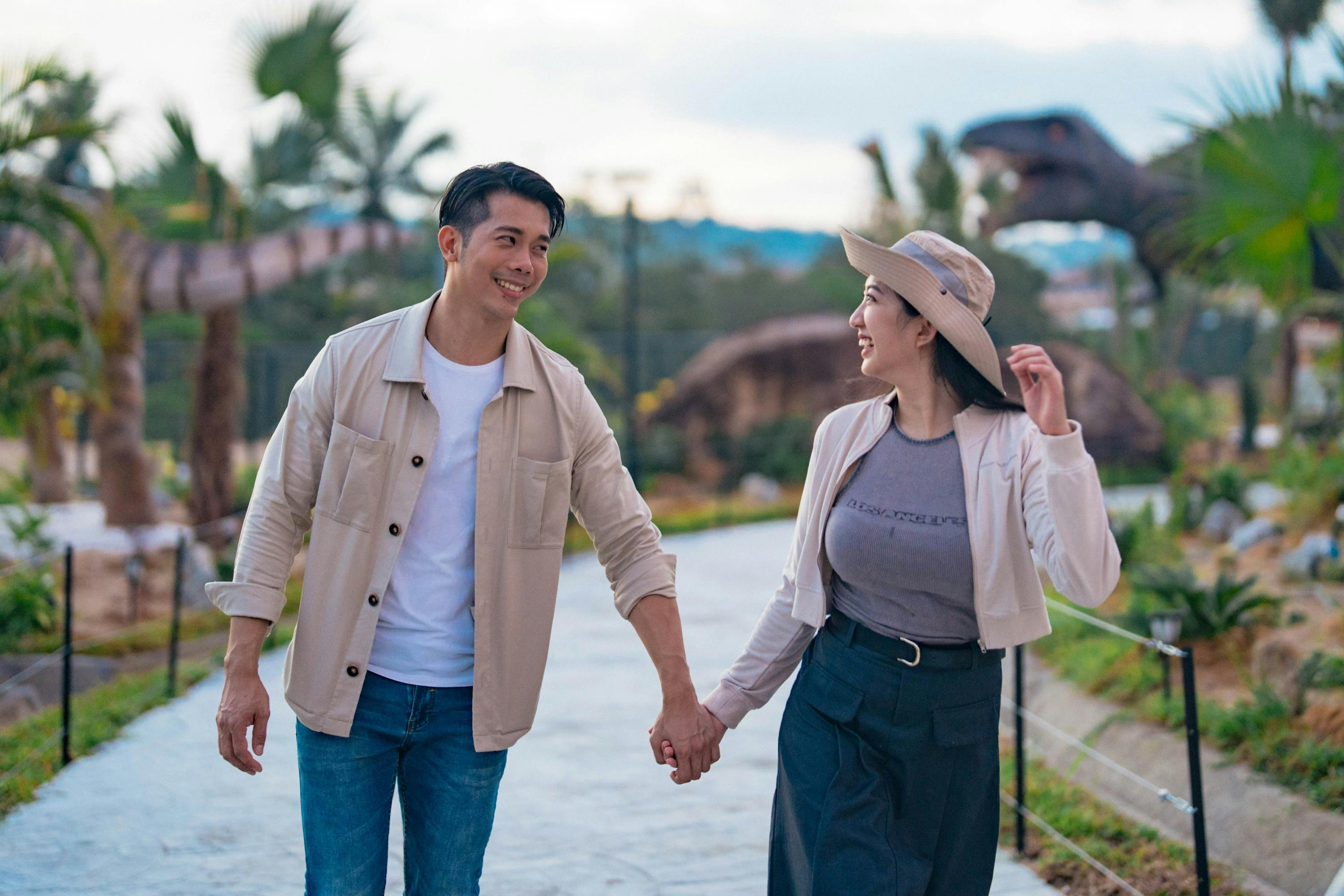 A couple holding hands and smiling at each other while walking outdoors on a pathway with greenery in the background.