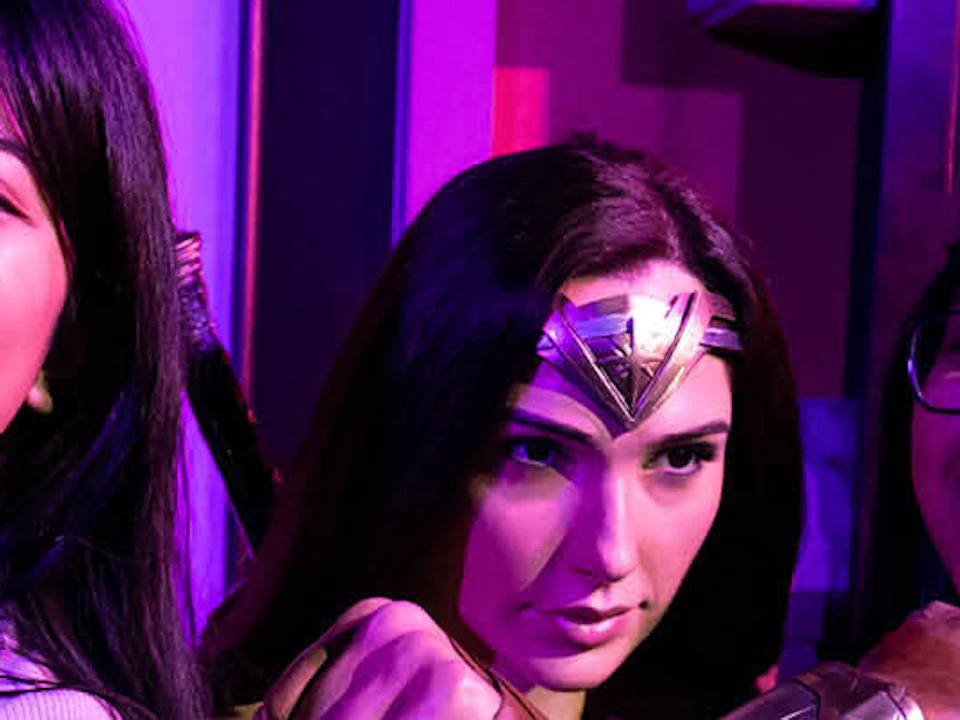 Two smiling women pose beside a Wonder Woman statue inside a room with vibrant lighting.