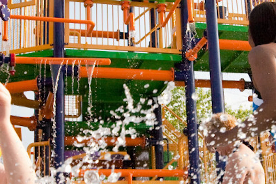 Children playing and splashing in a water park with colorful structures and slides in the background.