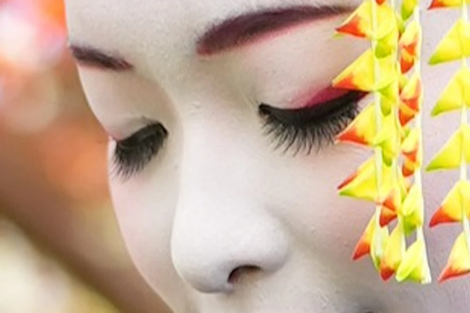 A person in traditional Japanese attire with white face makeup stands among vibrant autumn foliage.