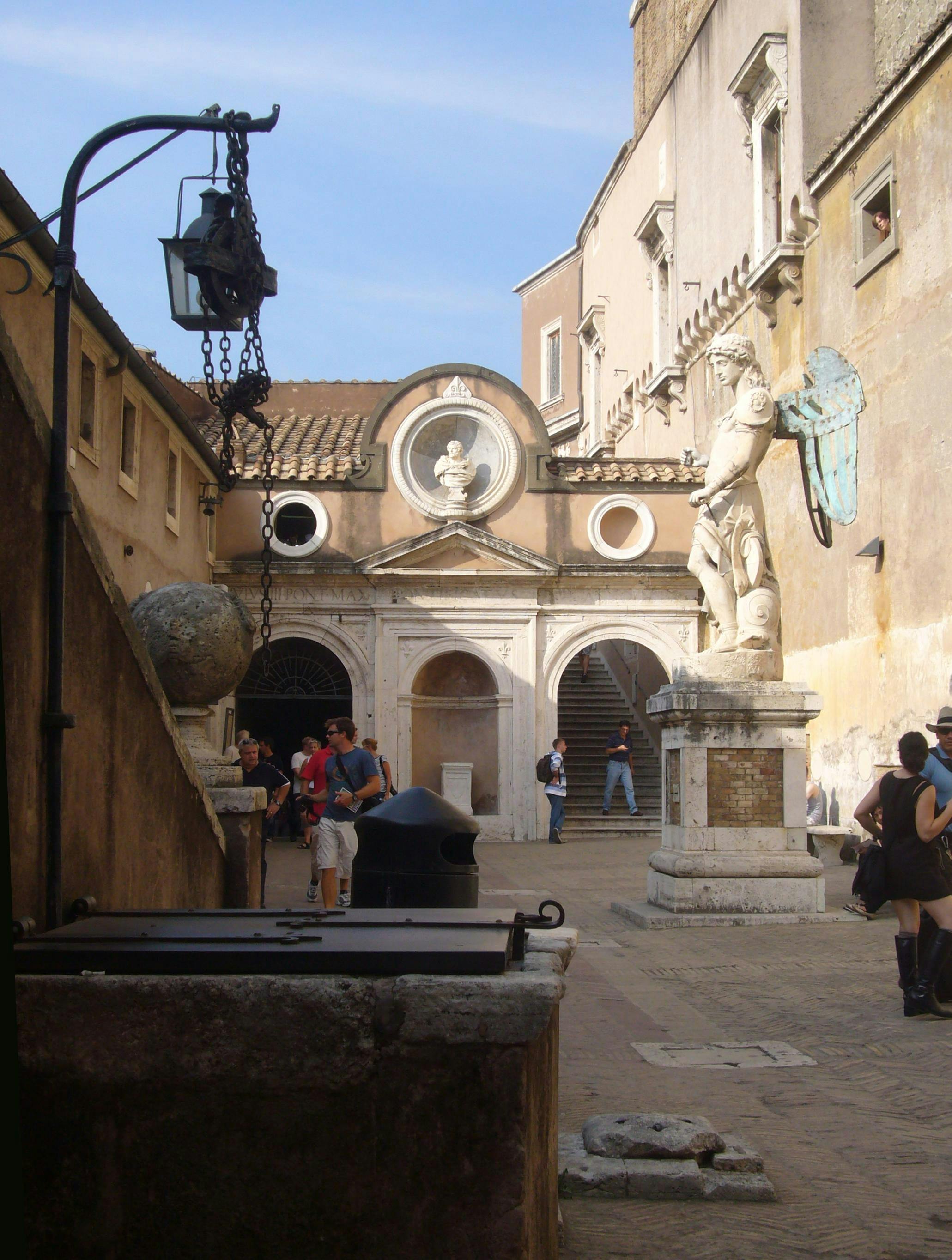 Двор с каменной статуей, люди ходят, арочные дверные проемы и лестница, ведущая к зданию, увенчанному бюстом.