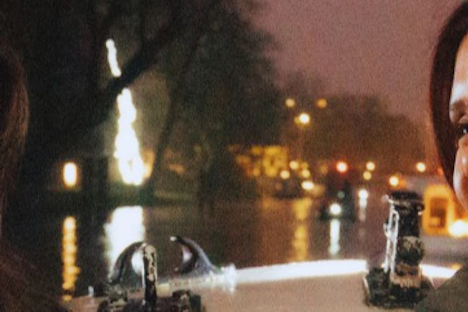 Two women smiling at each other, holding coffee cups, outside at night with trees and lights in the background.