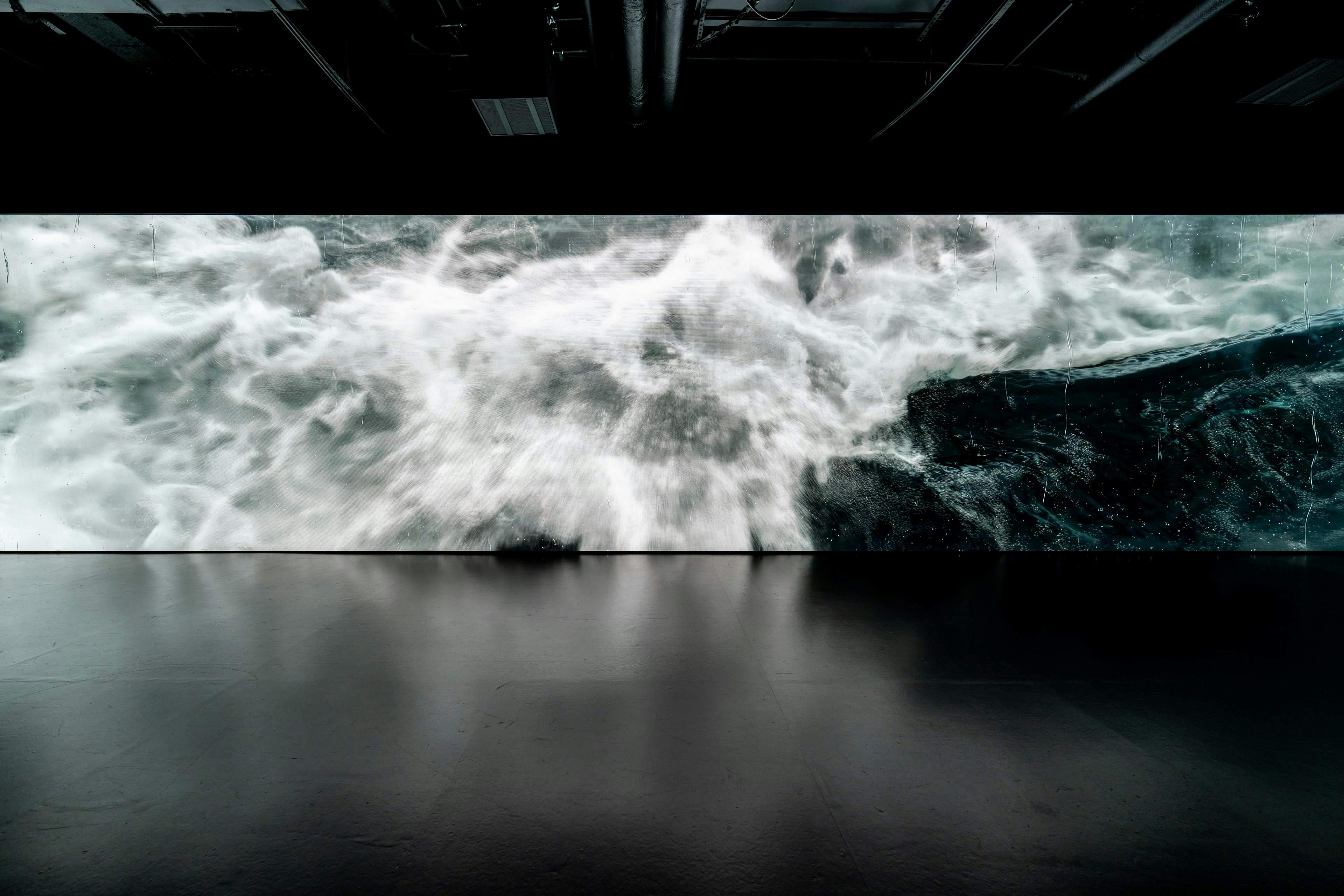A large digital screen displays a turbulent, white and gray swirling pattern, likely simulating a stormy sea, in a dark room.