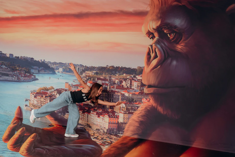A woman poses playfully as if being held by a large King Kong figure against a scenic city and sunset backdrop.