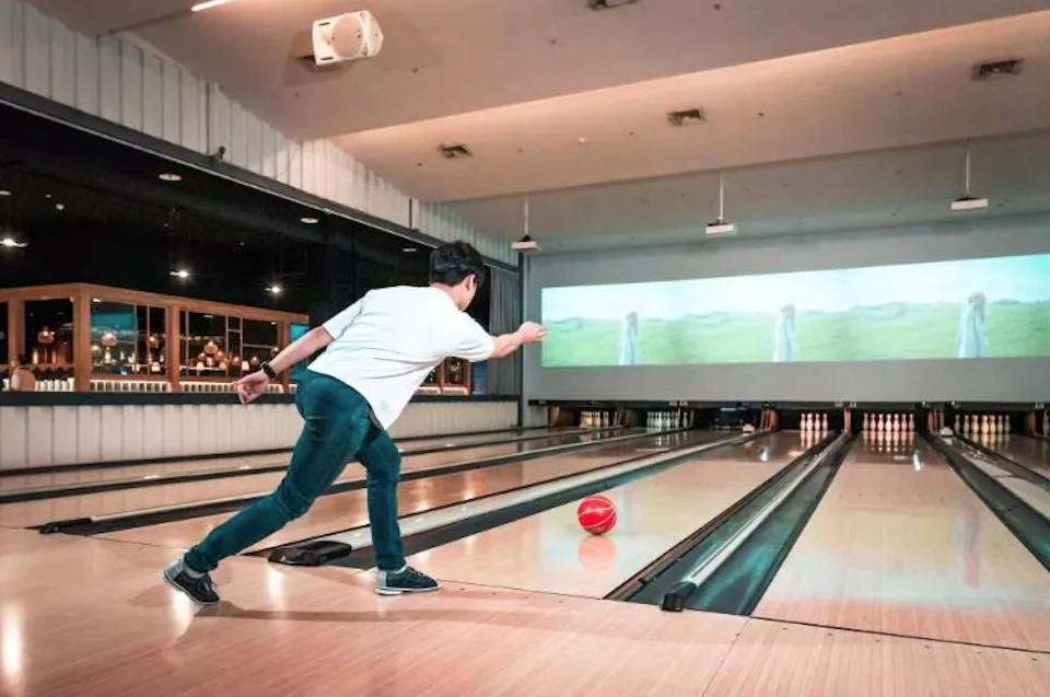 A person is bowling in an indoor alley, releasing a red ball towards the pins. A screen is visible above the lanes.
