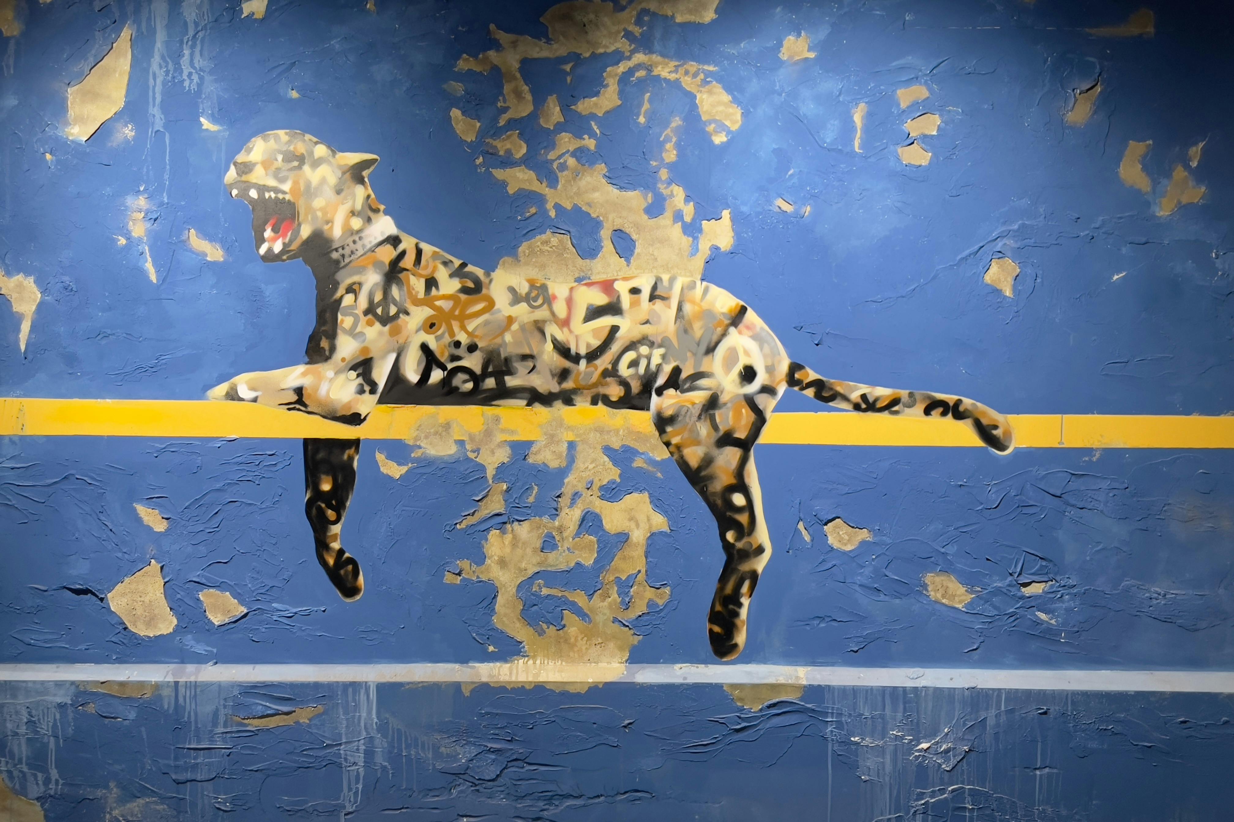 Fototapet i graffitistil av en rytande leopard med ett kollage av taggar på kroppen, uppflugen på en gul linje mot en blå, nödställd vägg.