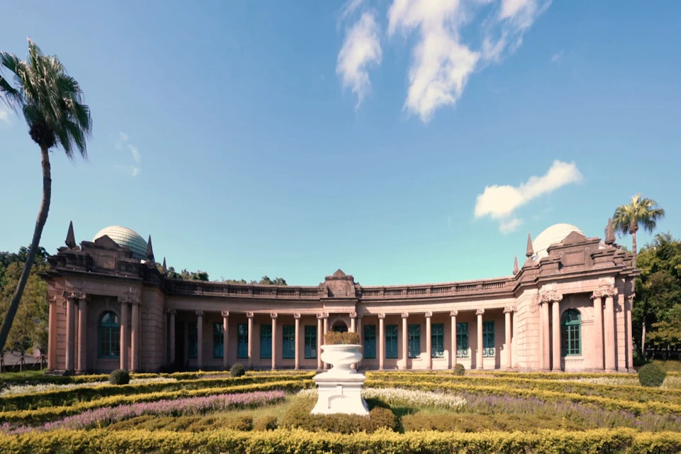 A neoclassical building with columns and domed ends, surrounded by manicured gardens under a blue sky with clouds.