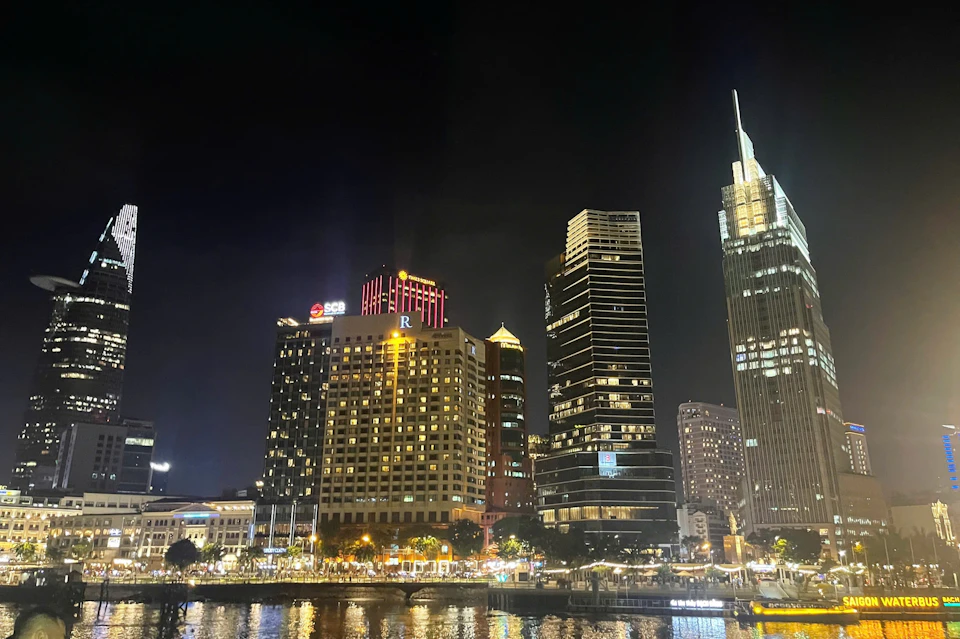 Night skyline featuring illuminated high-rise buildings, including a red-lit "R" building, reflecting on a calm waterfront.