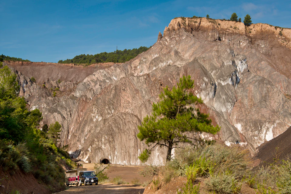 A rugged landscape featuring a large rock mountain, tunnel entrance, green trees, and a parked vehicle under a clear blue sky.