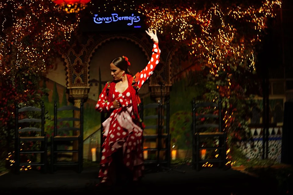 A person in a red polka dot dress performs a Flamenco dance on stage, adorned with lights and chairs, at "Torres Bermejas."