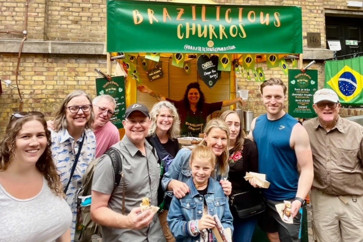 Μια ομάδα ανθρώπων ποζάρει μπροστά από ένα περίπτερο με ένα πράσινο πανό που γράφει "Βραζιλιάνικα Τσούρος".