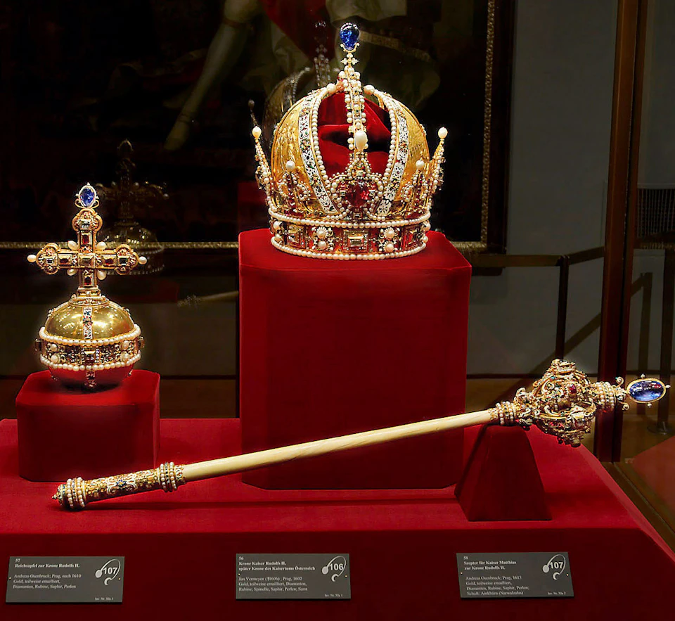 A golden crown, orb, and scepter adorned with jewels displayed on red velvet stands in a museum exhibit.