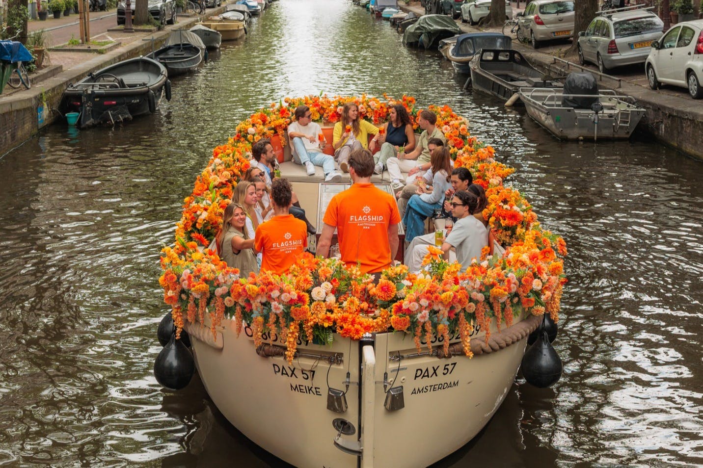 Ein mit orangefarbenen Blumen geschmücktes Boot befördert eine Gruppe von Menschen auf einem Kanal, an dessen Ufern geparkte Autos stehen.