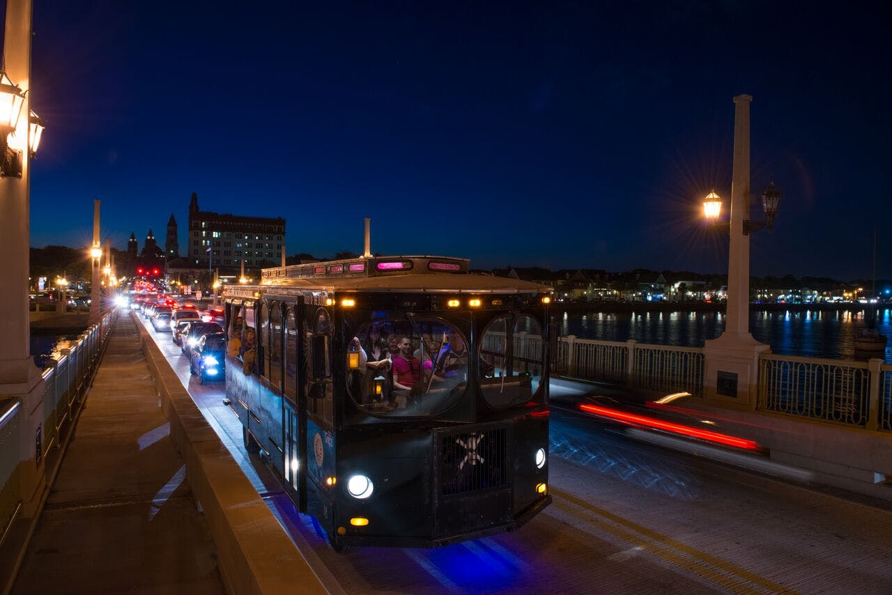 A trolley full of people travels over a bridge at night with city lights and traffic in the background.