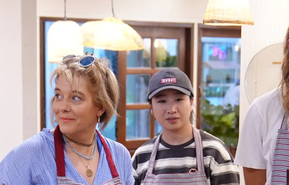 A group of people wearing aprons stand in a kitchen setting, with a cooking setup and an instructor demonstrating.