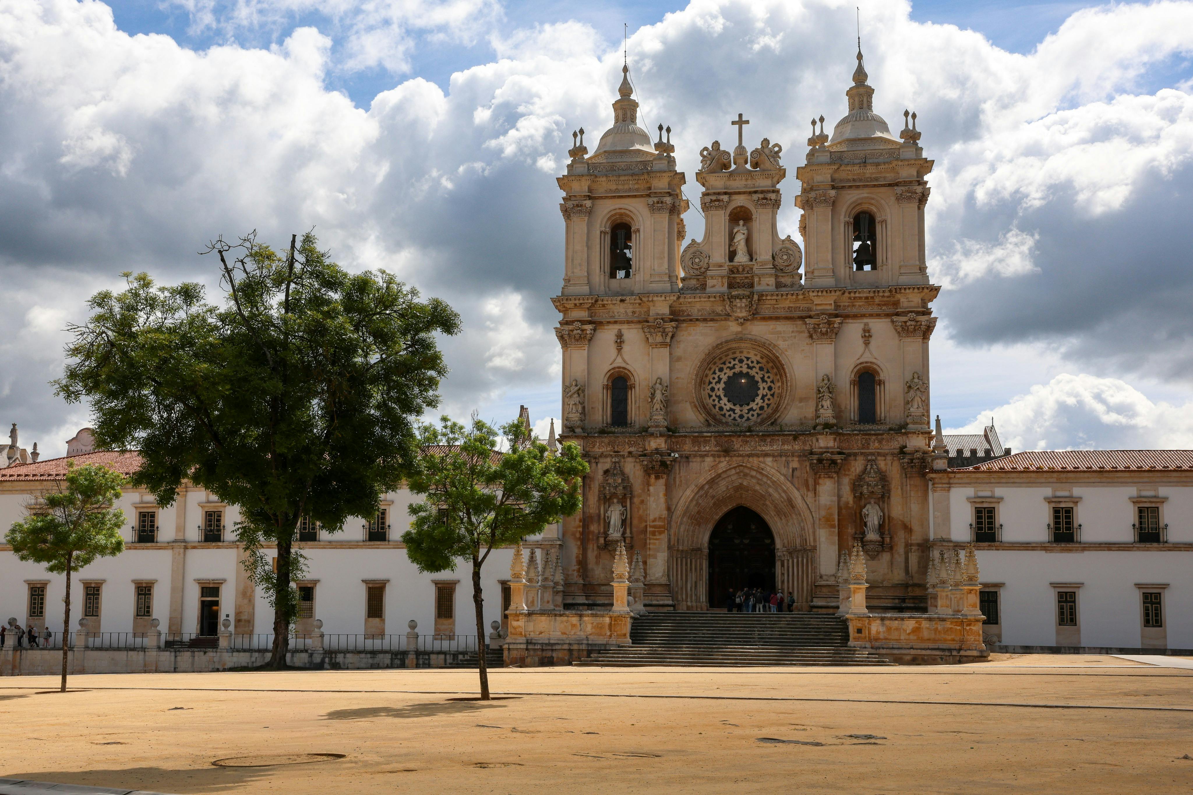 Eine historische Kirche mit zwei Türmen und komplizierten Details, vor einem wolkenverhangenen Himmel, mit Bäumen und einem Platz im Vordergrund.
