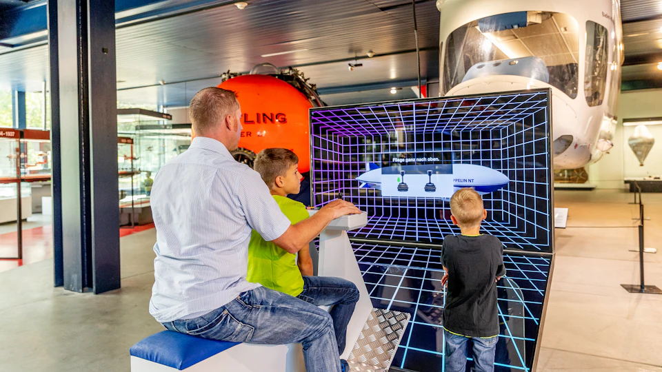 A man and two boys interact with a digital exhibit showcasing a grid and diagrams, with a large orange object in the background.