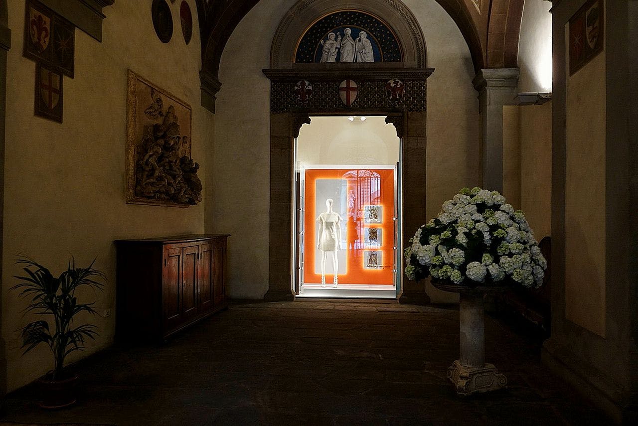 Dimly lit hallway featuring a mannequin in a glass case, a bouquet of white flowers, and an ornate arch with religious artwork.