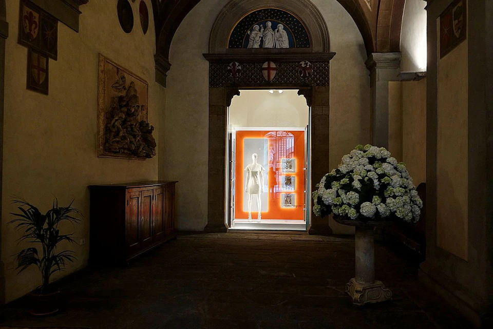 Dimly lit hallway featuring a mannequin in a glass case, a bouquet of white flowers, and an ornate arch with religious artwork.