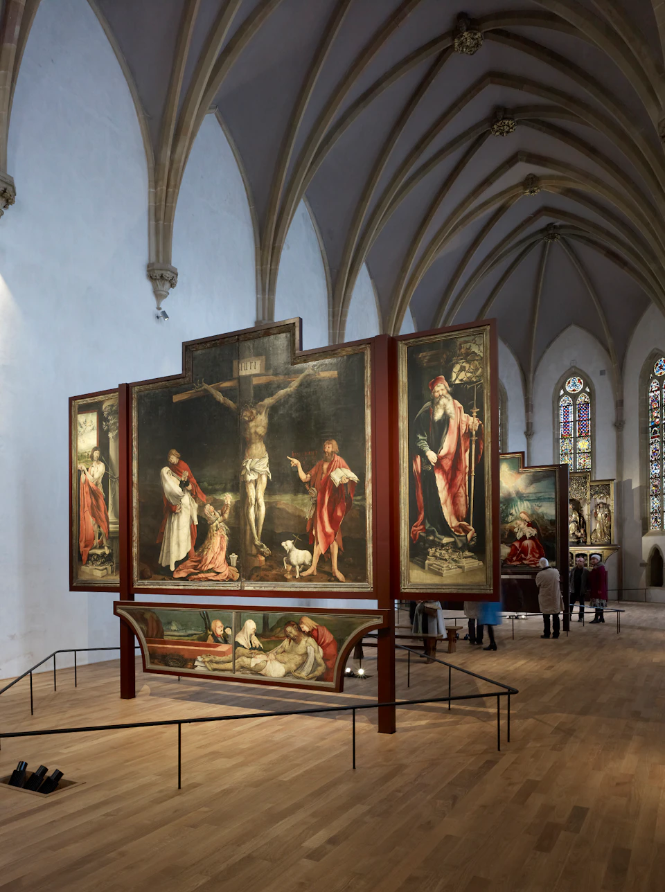 A cathedral interior showing ornate altarpieces with religious paintings and visitors observing the artwork.
