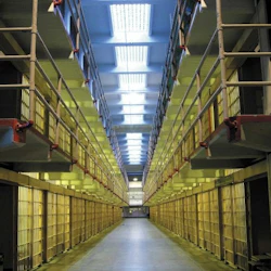 A symmetrical view of a prison hallway flanked by two levels of barred cells, lit by overhead fluorescent lights.