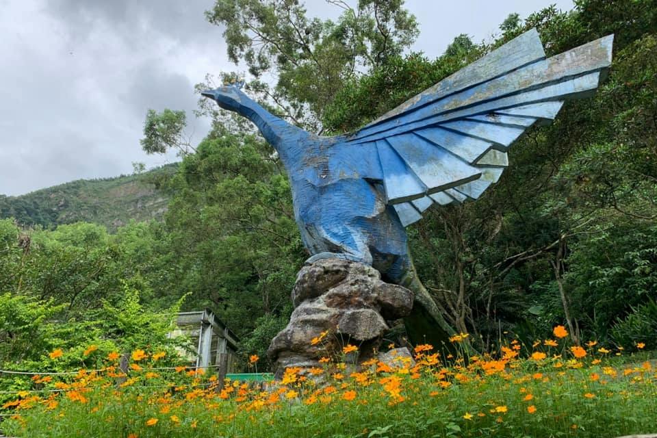 A large blue bird sculpture with extended wings stands on rocks surrounded by orange flowers and greenery.