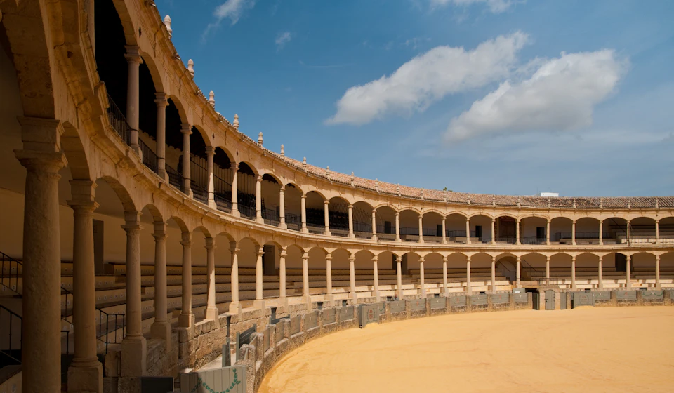 Curved, multi-tiered arched seating of an empty arena under a blue sky with scattered clouds.