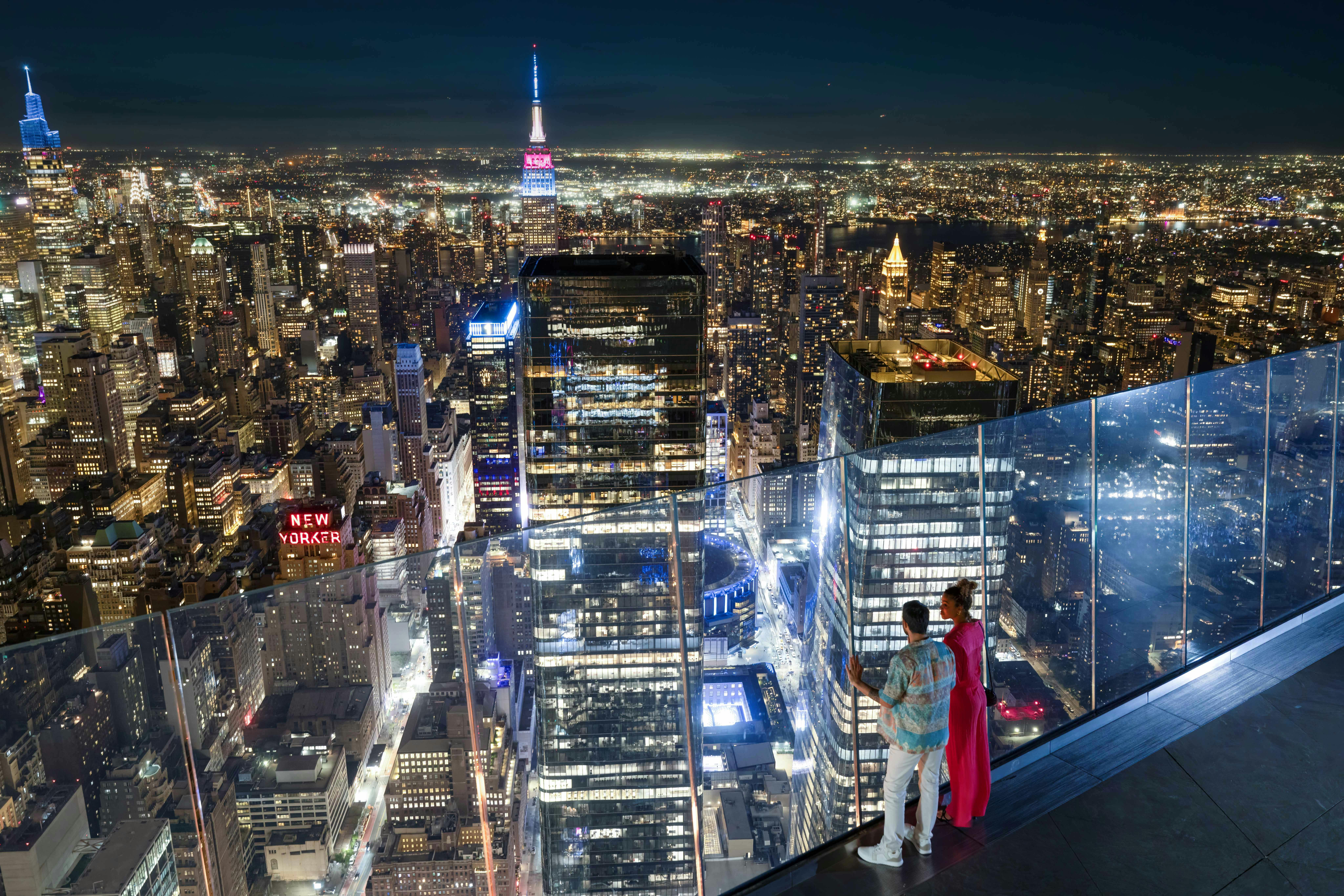 Due persone si trovano su un balcone di vetro che si affaccia su un paesaggio urbano illuminato di notte, con grattacieli e una torre prominente in vista.