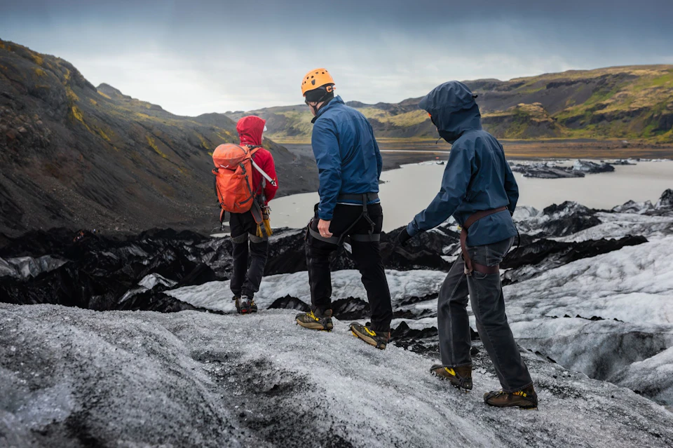 Three people in hiking gear and helmets walk on a glacier with a scenic mountainous background.