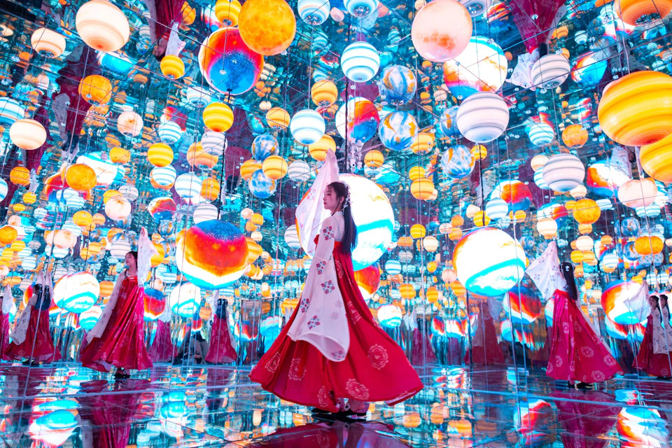 A woman in a vibrant traditional dress stands in a mirrored room filled with colorful hanging spheres resembling planets.