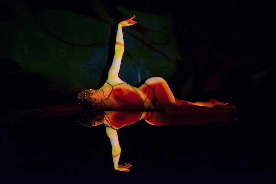 A performer is posing in a colorful, illuminated outfit, reflecting on a shiny surface against a dark background.