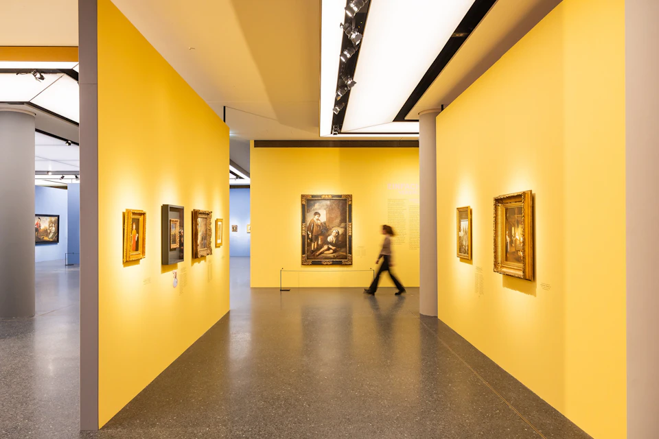 A person walks through an art gallery with yellow walls displaying framed paintings under bright lighting.