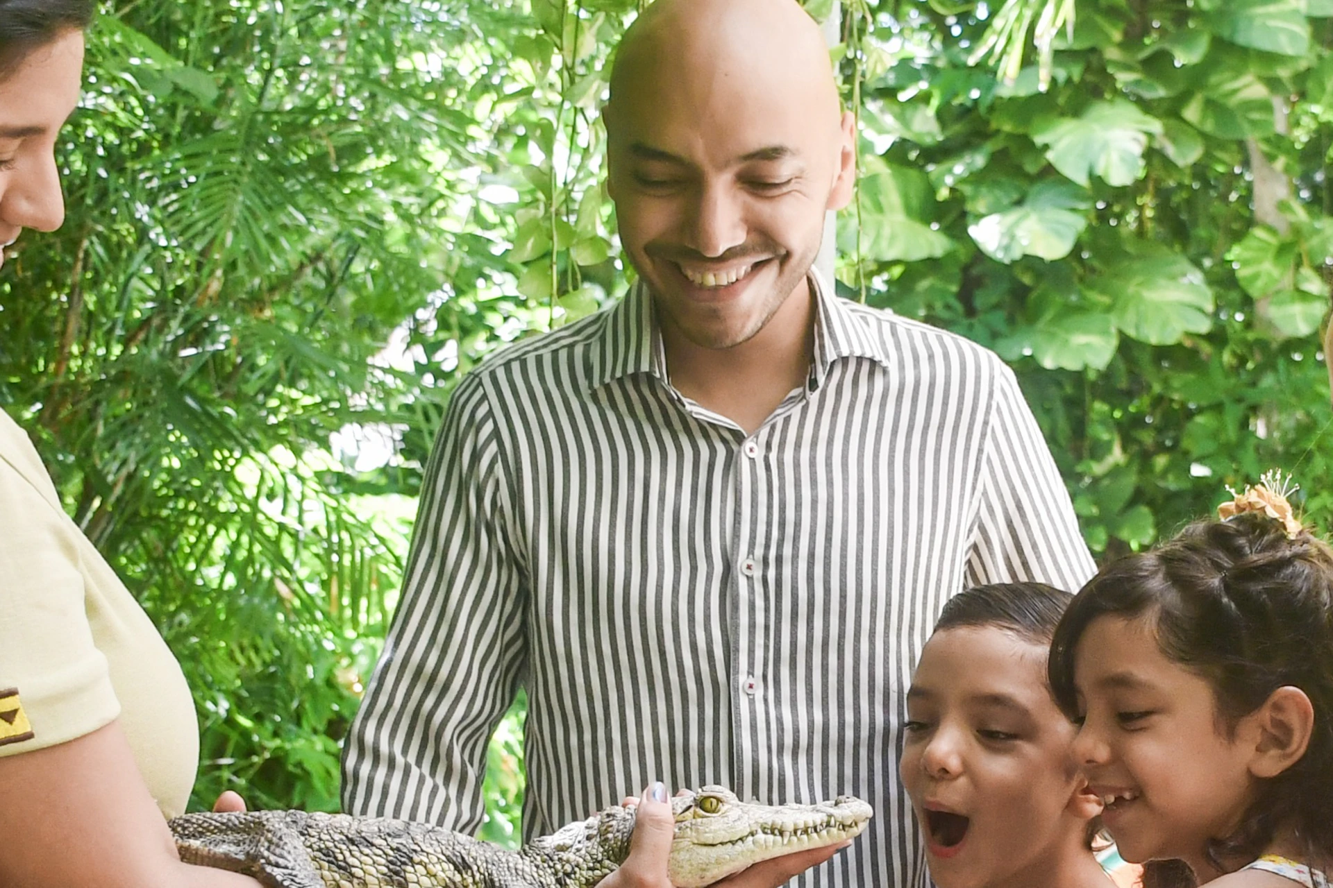 A zookeeper shows a small crocodile to a smiling family, including two children, in a lush, green setting.