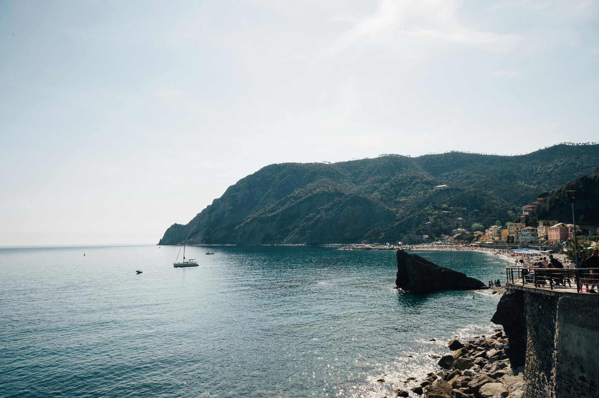 宁静的滨海风光，晴朗的天空下有平静的蓝色海水、帆船、岩石海岸和绿色的山丘。