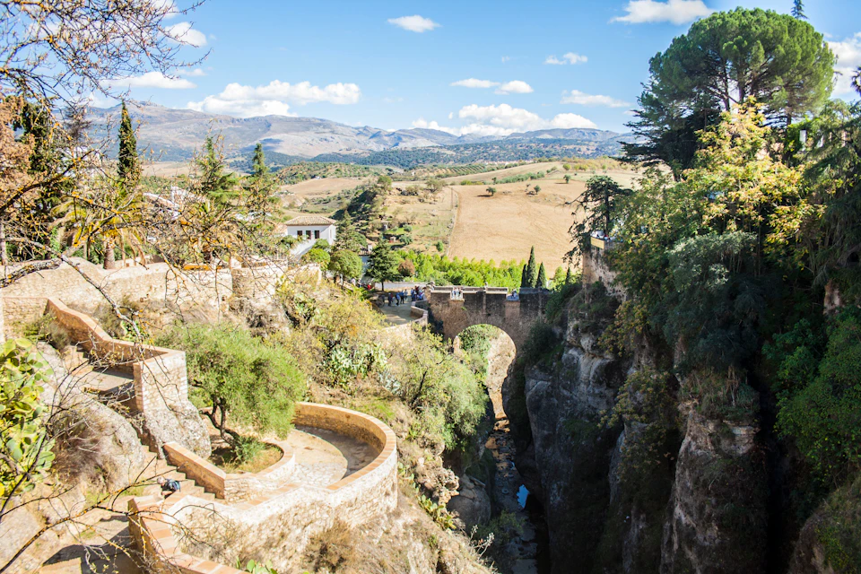 A stone bridge spans a deep gorge with lush vegetation, overlooking a rural landscape under a blue sky with scattered clouds.