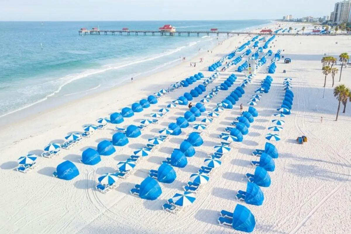A beach with rows of blue umbrellas and chairs, a pier extending into the ocean, and a clear sky above.