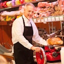 Un uomo sorridente con un grembiule aziona un'affettatrice in una gastronomia, circondato da varie carni e prodotti stagionati.