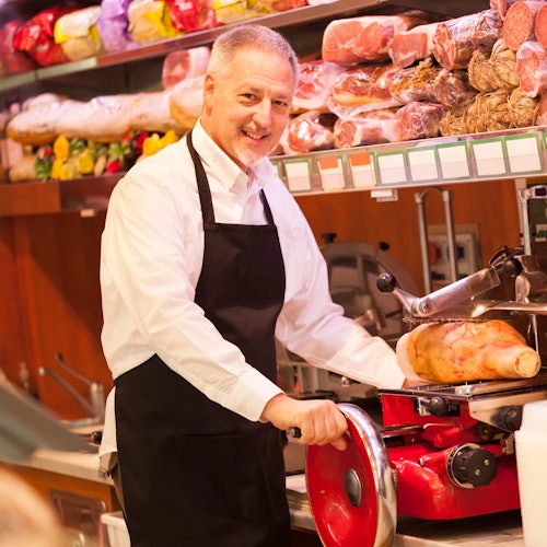 Un uomo sorridente con un grembiule aziona un'affettatrice in una gastronomia, circondato da varie carni e prodotti stagionati.