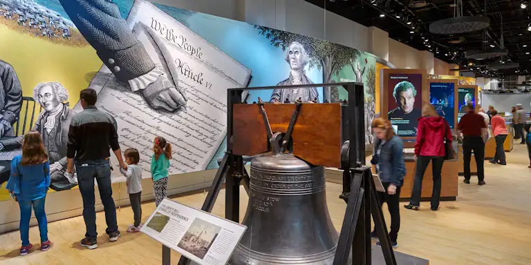 Osoby eksplorujące interaktywną wystawę historyczną z dużym dzwonkiem, kolorowymi malowidłami ściennymi i ekspozycjami informacyjnymi.