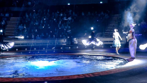 Performers in elaborate costumes dance near an illuminated circular water feature with a seated audience in the background.