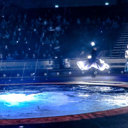 Performers in elaborate costumes dance near an illuminated circular water feature with a seated audience in the background.