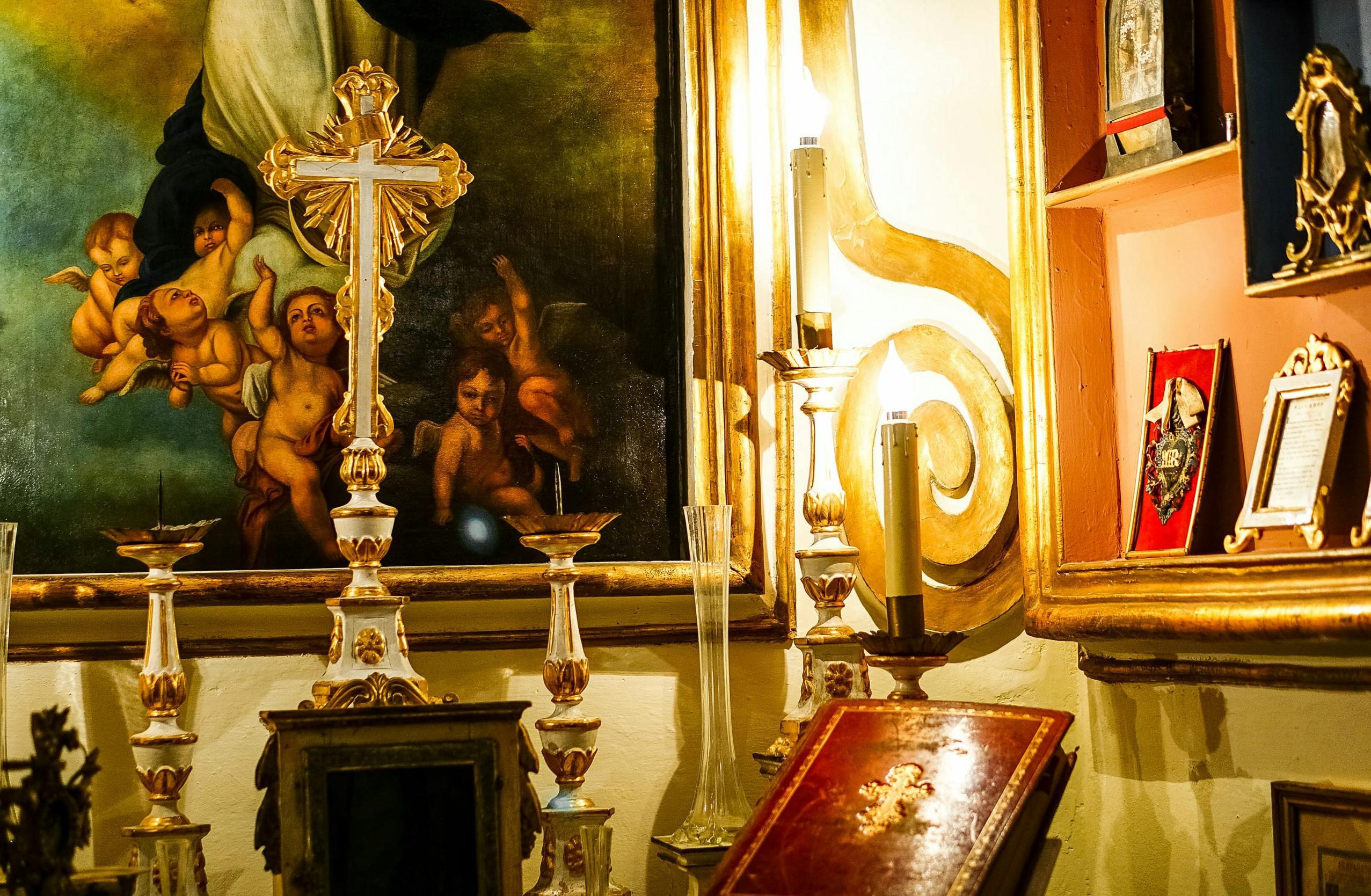 Et udsmykket kors og lysestager foran et religiøst maleri, der forestiller engle og en klædt figur.