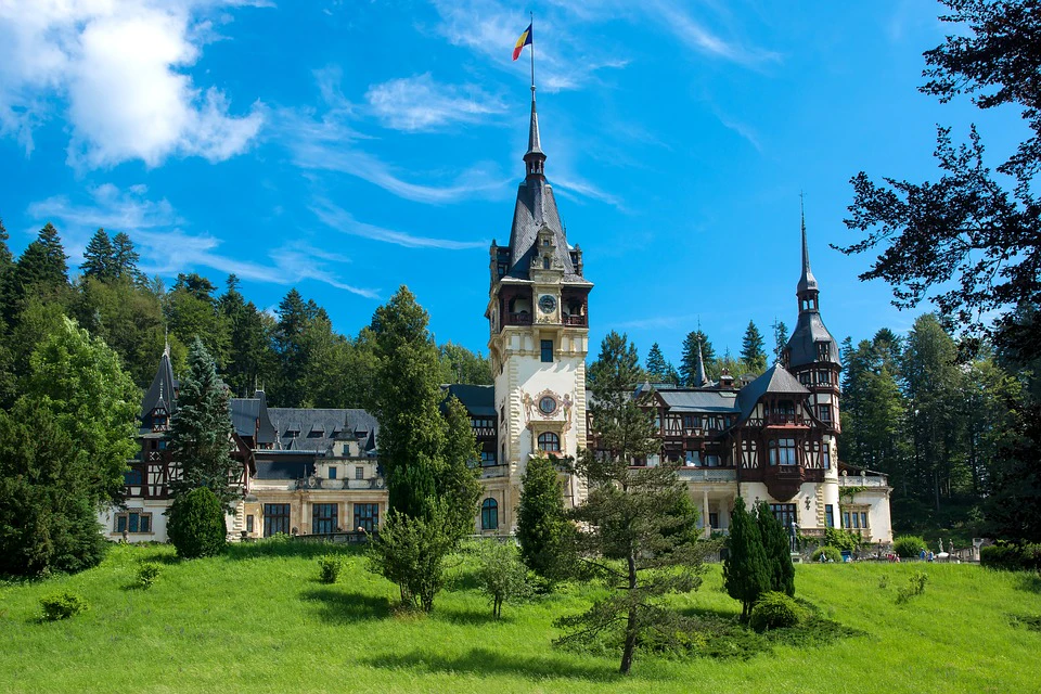 A picturesque castle with a tall spire and Romanian flag, surrounded by lush greenery and a clear blue sky.