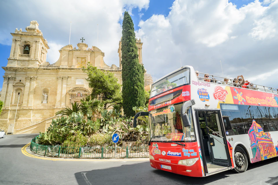 A red double-decker sightseeing bus in front of a historical building with a large tree and greenery.