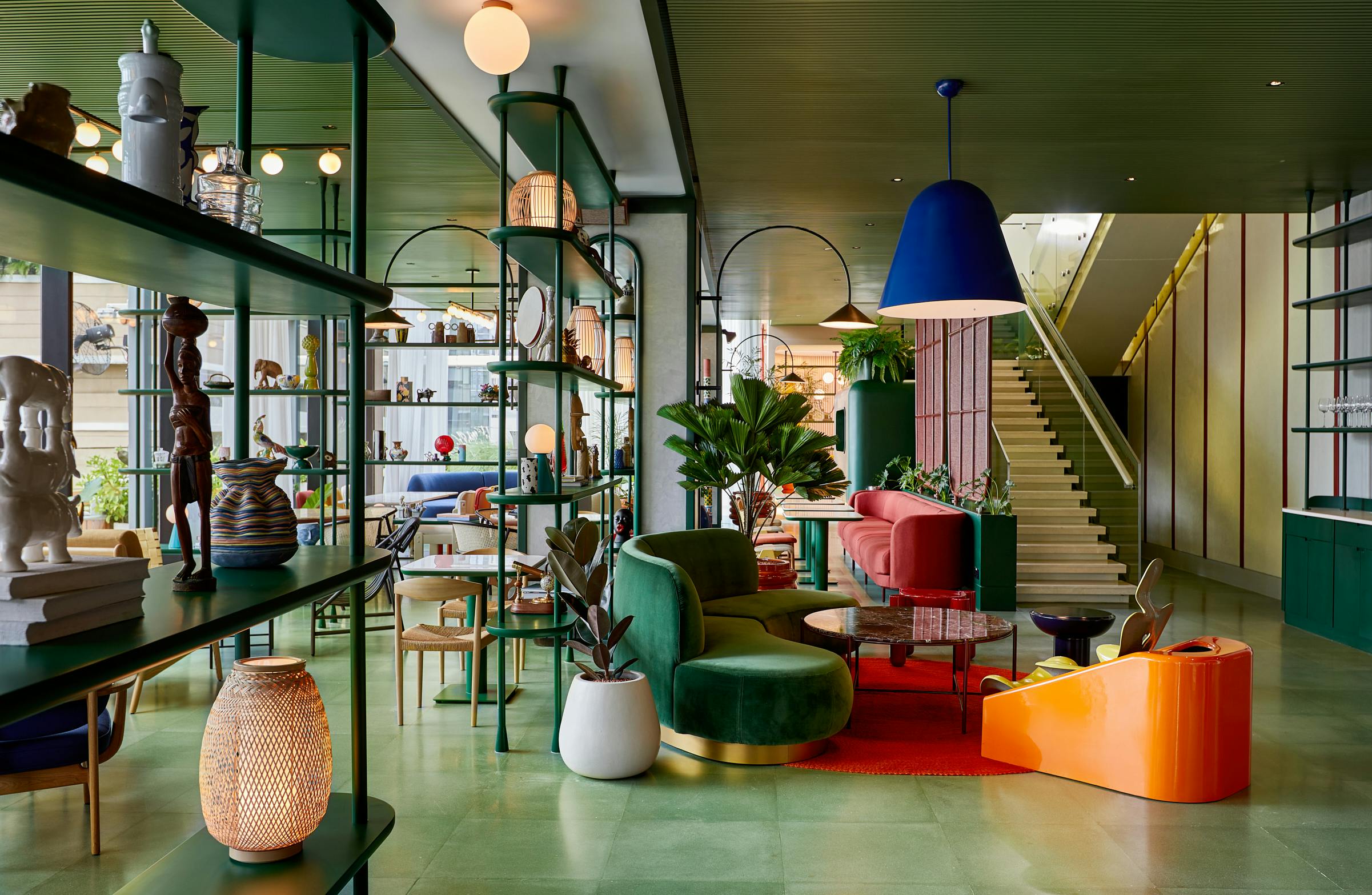 A colorful modern interior with green and red furniture, plants, shelves, and a staircase in the background.