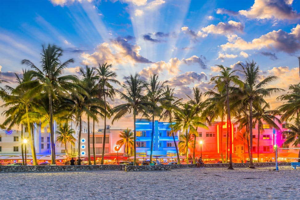 Palm trees in front of brightly lit buildings with neon lights at sunset, under a partly cloudy sky with sun rays.