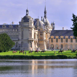 Ryttare till häst och människor som går framför ett stort slott med en damm i förgrunden och träd på vardera sidan.