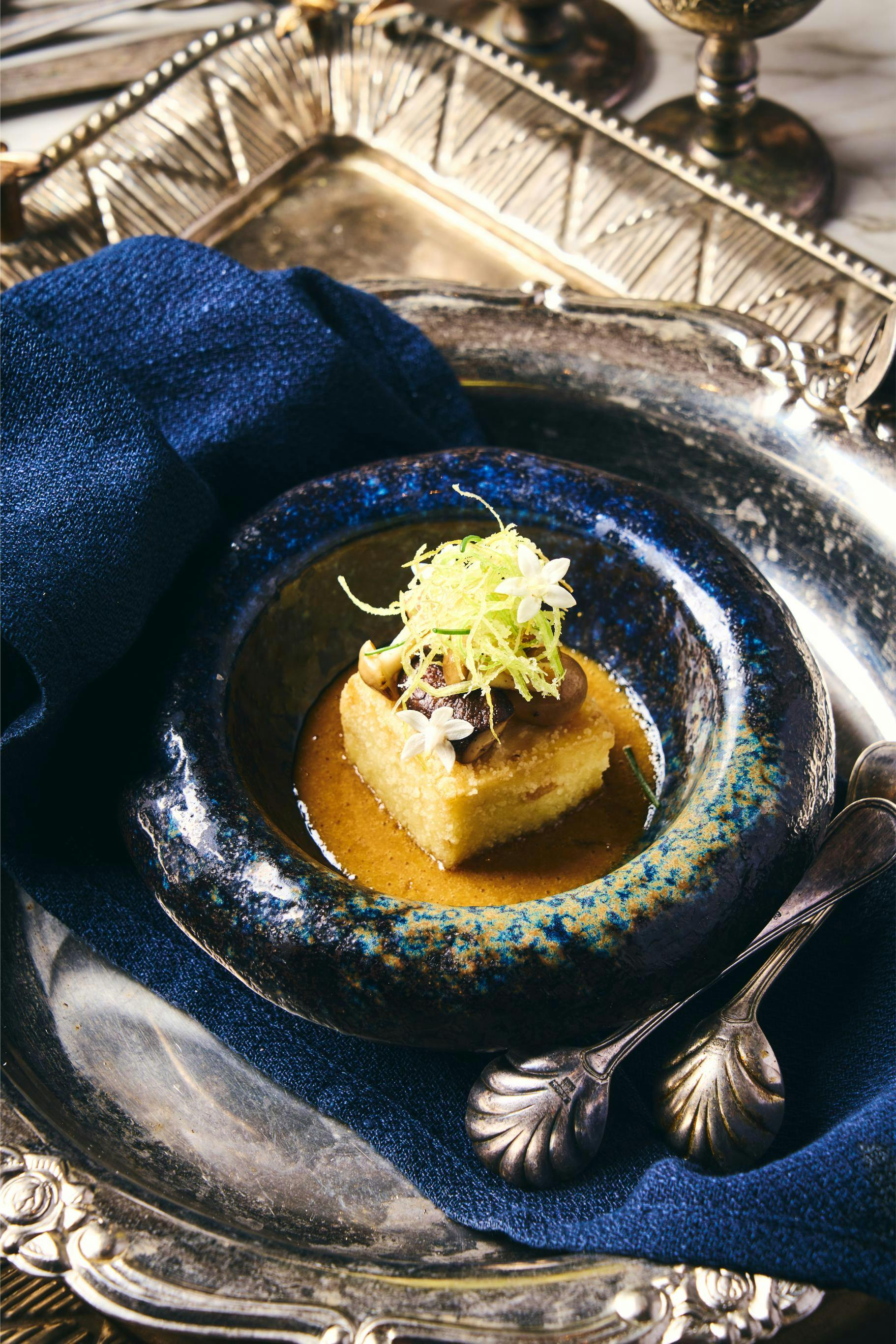 A plated dessert featuring a square cake topped with cream, green garnish, and flowers, served in a dark decorative bowl.