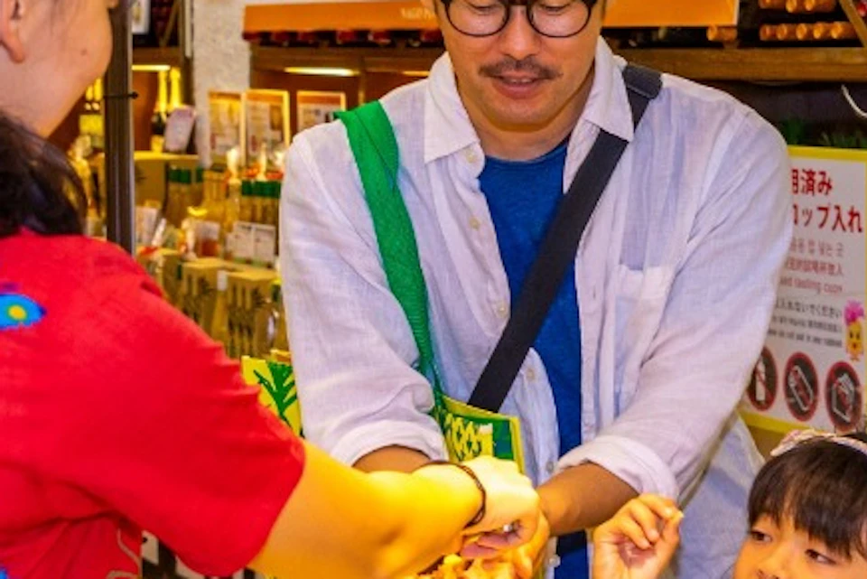 A shop employee in a red shirt assists a family with a young child in a store with yellow-packaged products on display.
